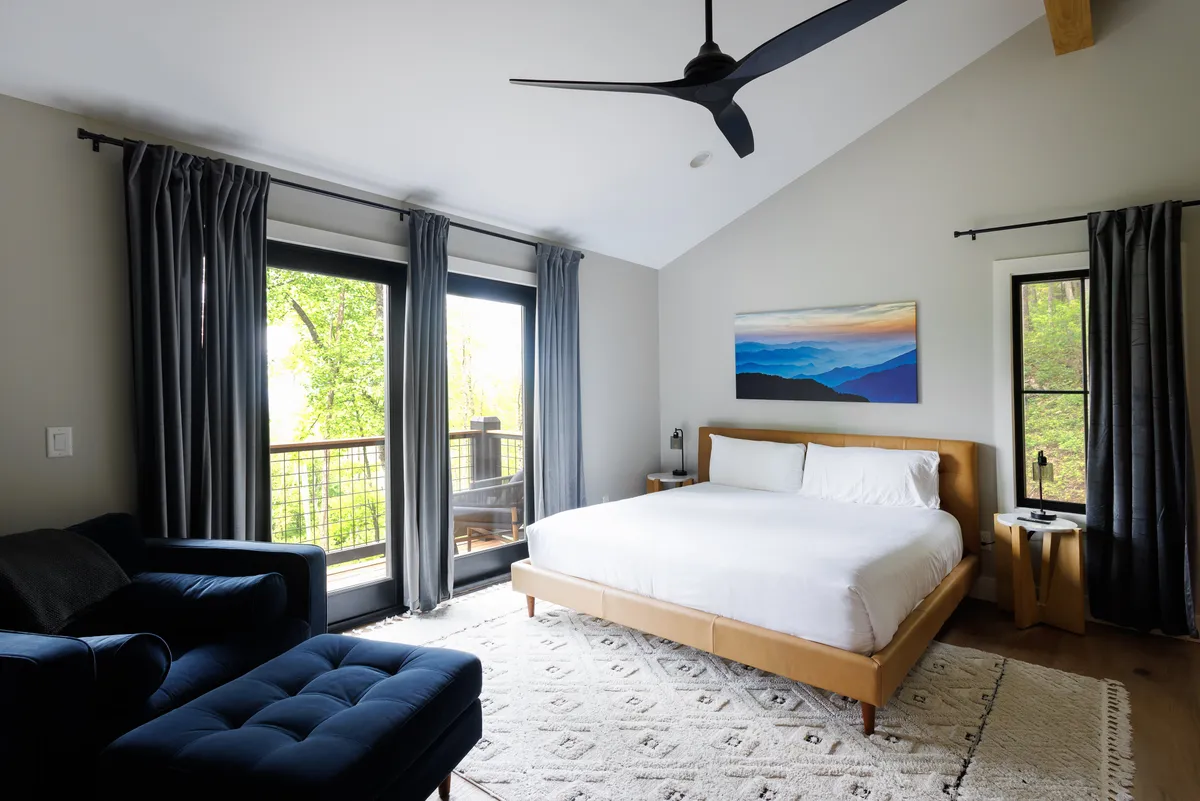 This image features a king-sized bed set against large windows that frame a serene mountain landscape painting, complemented by a blue accent chair. Soft natural light fills the room, while the inviting chair and calming artwork enhance the tranquil ambiance. The space exudes peacefulness and comfort, ideal for restful nights and quiet mornings.