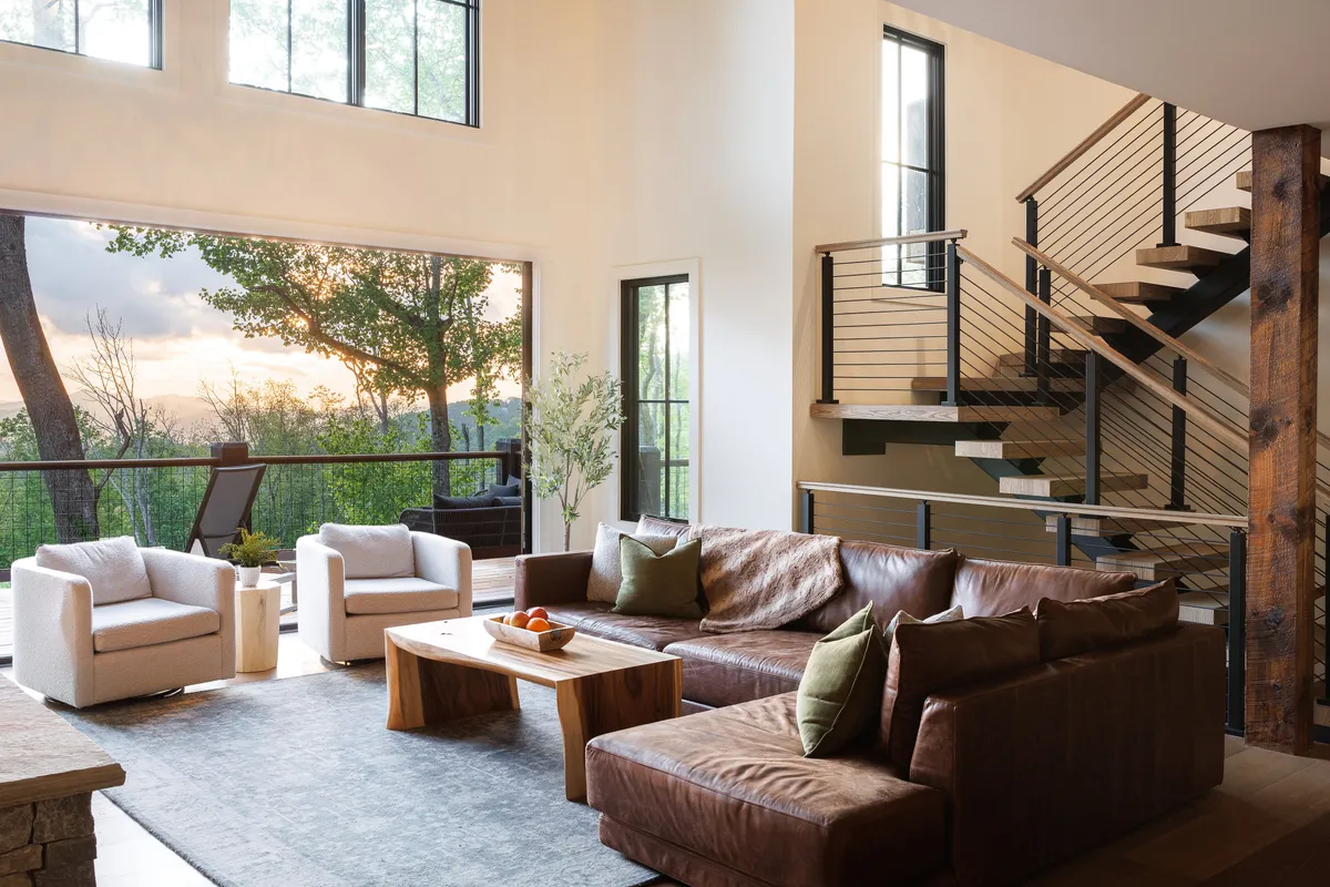 This image presents a warm, inviting living area featuring a plush leather sofa, a wood coffee table, and expansive windows that frame a glorious sunset over the mountains. The staircase adds architectural interest, and the overall mood is serene and welcoming, emphasizing connection and comfort.