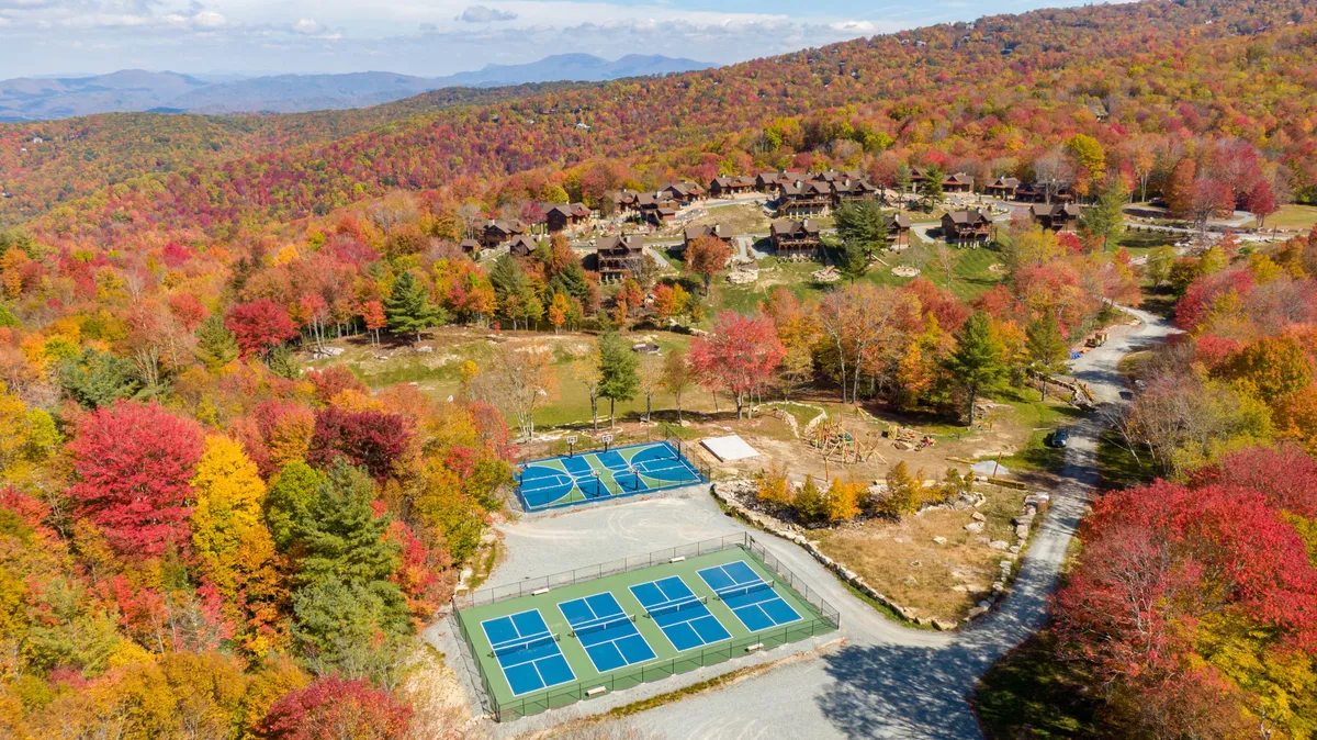 This image depicts vibrant autumn foliage surrounding tennis courts and rustic cabins, evoking a lively, active community environment. The bright leaves, open spaces, and visible recreational amenities create a cheerful, inviting atmosphere that signals year-round outdoor fun and seasonal beauty.