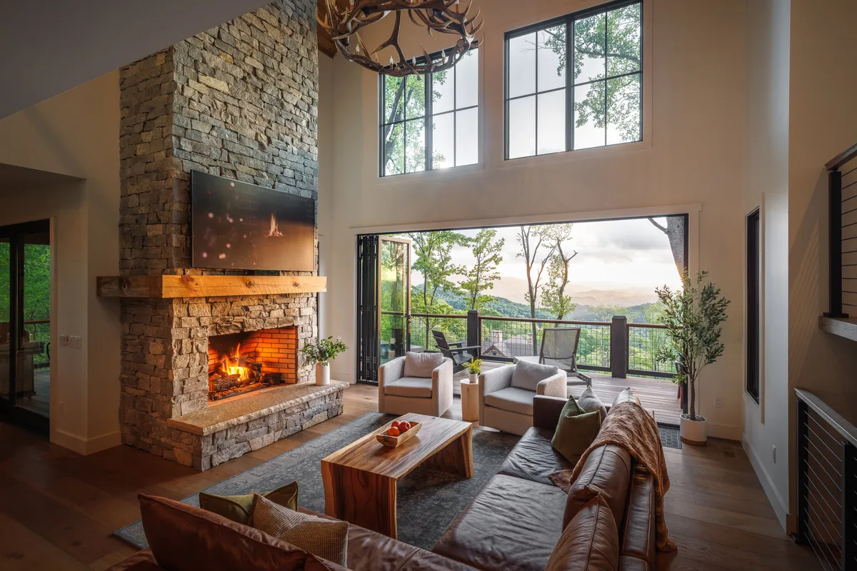 The image showcases a warm, inviting living room with a grand stone fireplace as the centerpiece, surrounded by plush leather seating and a rustic wooden coffee table. Floor-to-ceiling windows frame breathtaking sunset views over the mountains, filling the space with golden light and offering a perfect spot to gather and unwind.