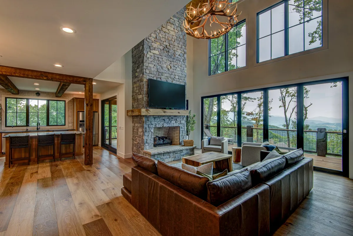 Showcasing a modern, spacious living room with a grand stone fireplace, large windows offering layered mountain vistas, comfortable seating, and a dramatic chandelier, this image captures both sophistication and the warmth of communal gathering.
