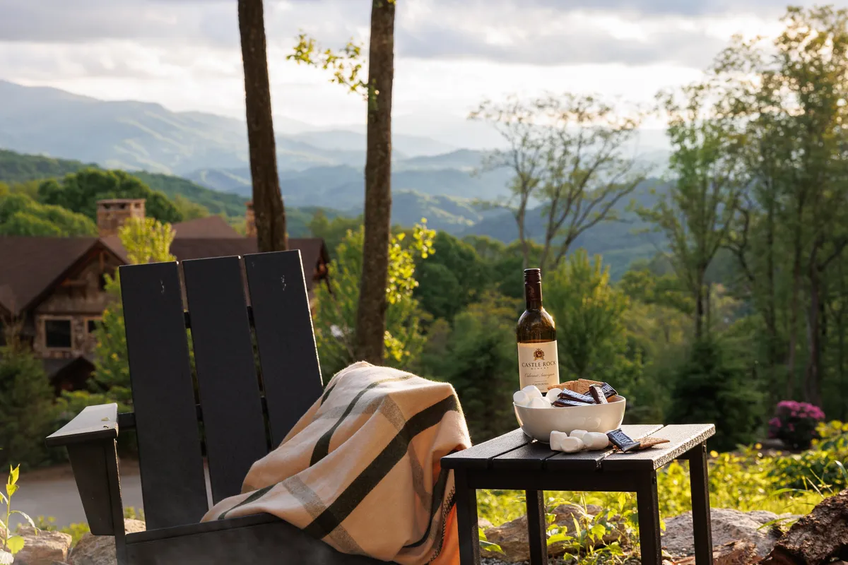 A cozy exterior setting featuring a chair draped with a blanket, a wine bottle, and s'mores ingredients, all set against a dramatic mountain sunset. The arrangement suggests relaxation and togetherness while highlighting classic mountain evening activities like making s'mores around a fire.
