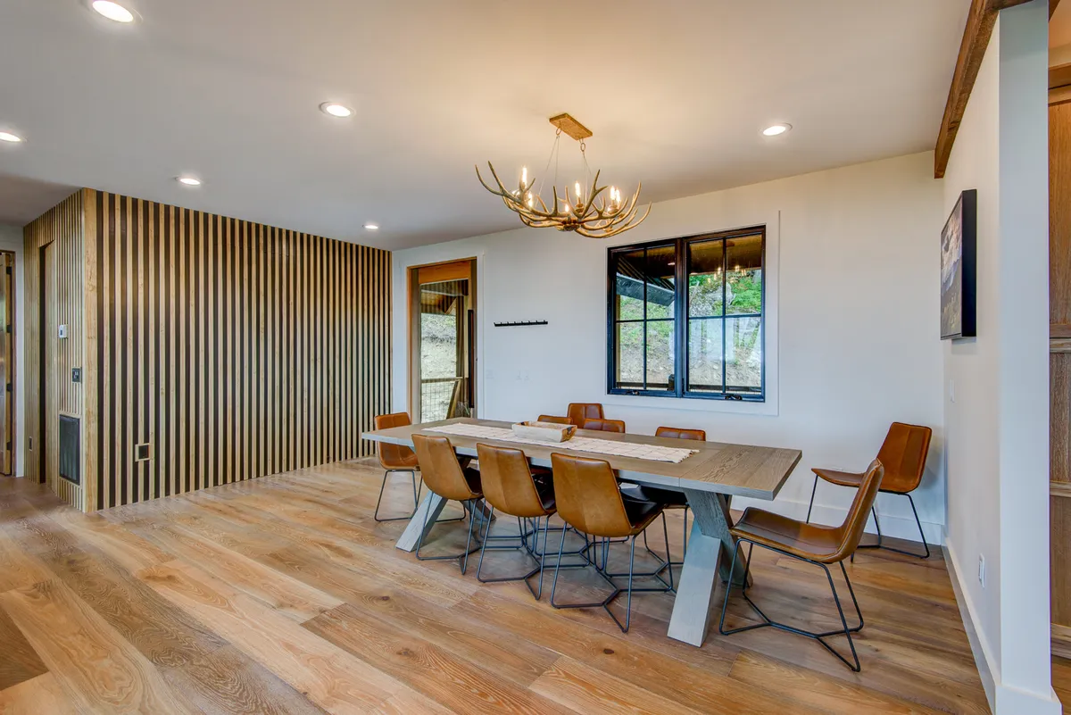 A modern, inviting dining area with an elegant table set beneath a statement chandelier, wooden accent wall, and large windows. The space is ready for family dinners, festive meals, or lively conversation, with a layout that encourages togetherness.