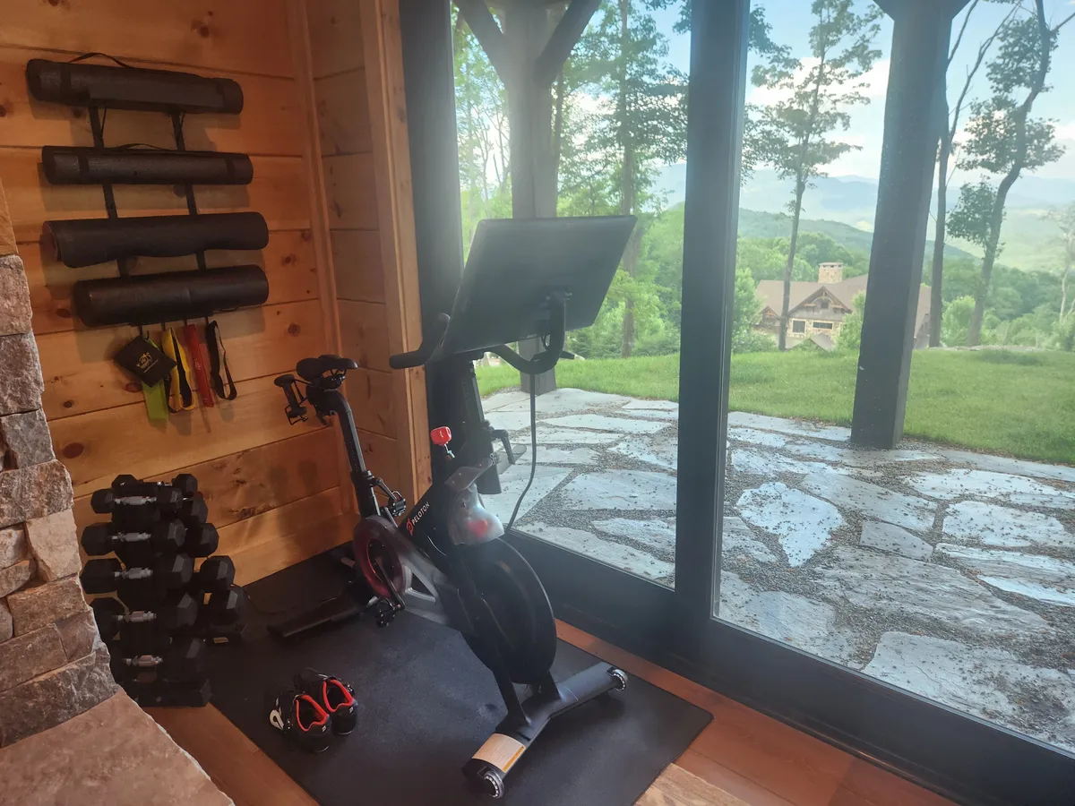 A bright and motivational private gym space equipped with an exercise bike, weights, and yoga mats. Large windows flood the area with natural light and offer a scenic mountain view, seamlessly integrating wellness with the beauty of the outdoors.