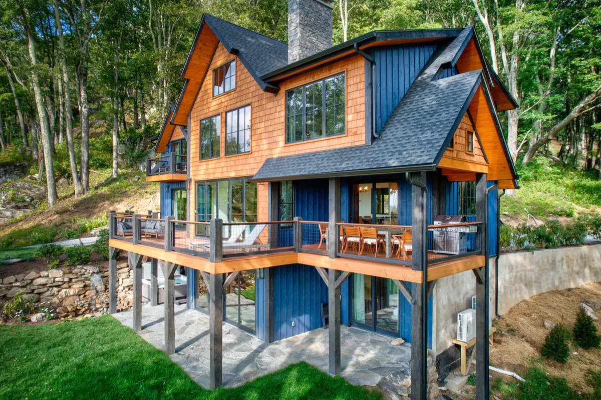 A welcoming exterior shot of the retreat, showcasing a spacious wooden deck, large windows, a stone chimney, and a blue and wooden facade. The deck is set up for outdoor dining and entertaining, with plenty of space for friends to gather and enjoy panoramic views.