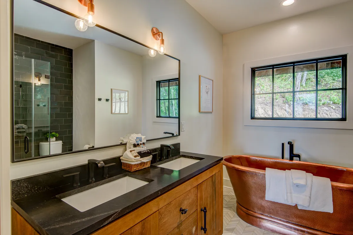 A luxurious en-suite bathroom with a freestanding copper bathtub, double sink vanity, large mirror, and windows letting in natural light. Decorative wall art adds an elegant touch, while the overall design is both tranquil and inviting—ideal for relaxation and self-care.