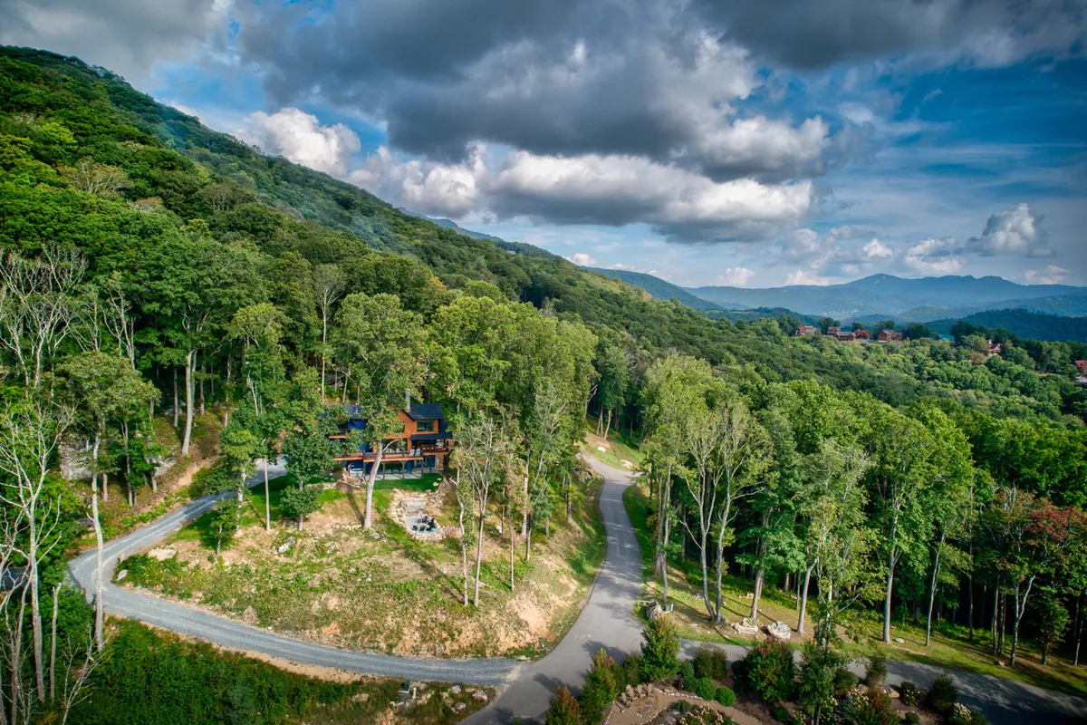 A view of the house nestled within a dense forest, with a winding road leading up to it and a backdrop of mountains. The surrounding trees and mountain views create an immersive sense of being in the heart of nature, inviting exploration and outdoor adventure.