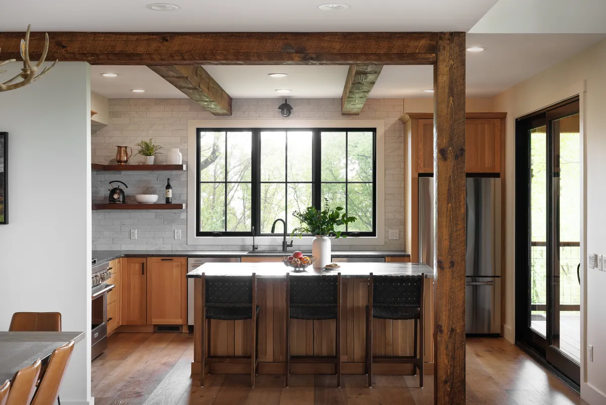 A spacious, stylish kitchen with a sizable island, large windows letting in abundant natural light, prominent wooden beams, and decorative shelving. The layout encourages both cooking and socializing, with enough space for guests to gather around.