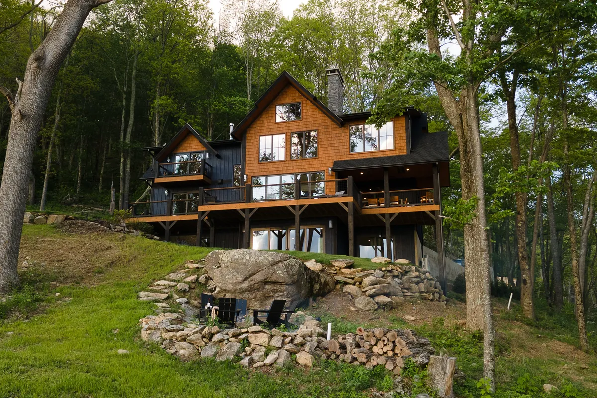The focus is on a large wooden deck with a stone fire pit, surrounded by trees and unique mountain architecture. The scene suggests communal gatherings—guests enjoying meals outdoors, sharing stories by the fire, or relaxing after a day of adventure, all with nature as the backdrop.