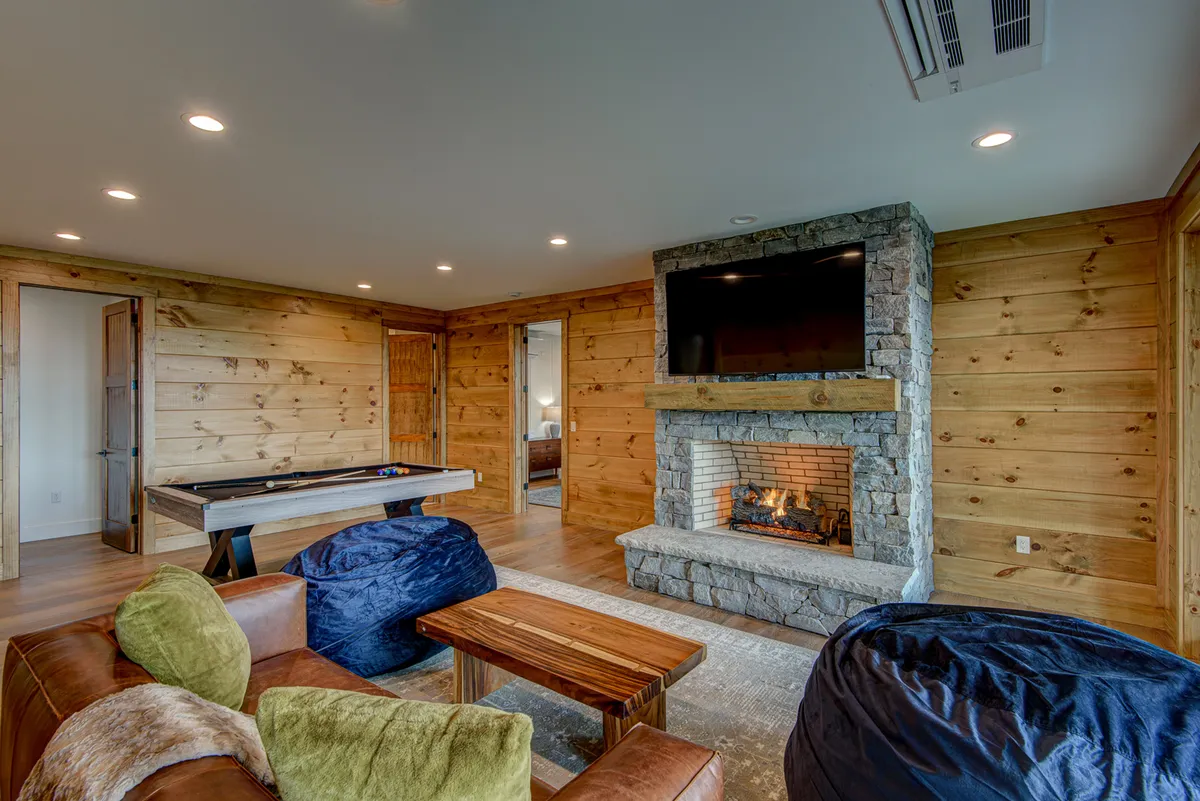 A cozy lower level living area featuring a fireplace, a modern pool table, and comfortable seating. The space is accented with wood paneling and soft lighting, creating a welcoming environment for both relaxation and playful competition. This room is designed for movie nights, social gatherings, and spirited games.