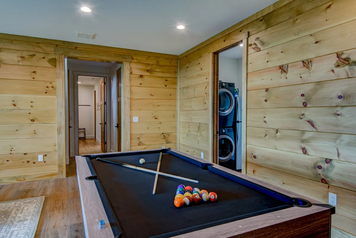 The interior game room showcases a pool table in a rustic space with wooden walls and doorways, inviting friendly competition and social interaction. The design is casual and relaxed, ideal for family game nights and laughter-filled gatherings.