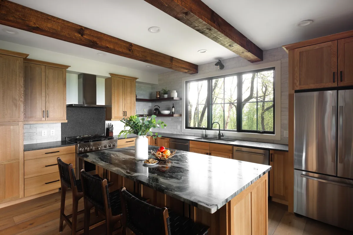 A warm, welcoming kitchen with a central island featuring a marble countertop, large window with mountain views, and exposed wooden beams overhead. The space is staged for cooking and gathering, with ample natural light enhancing the inviting atmosphere.