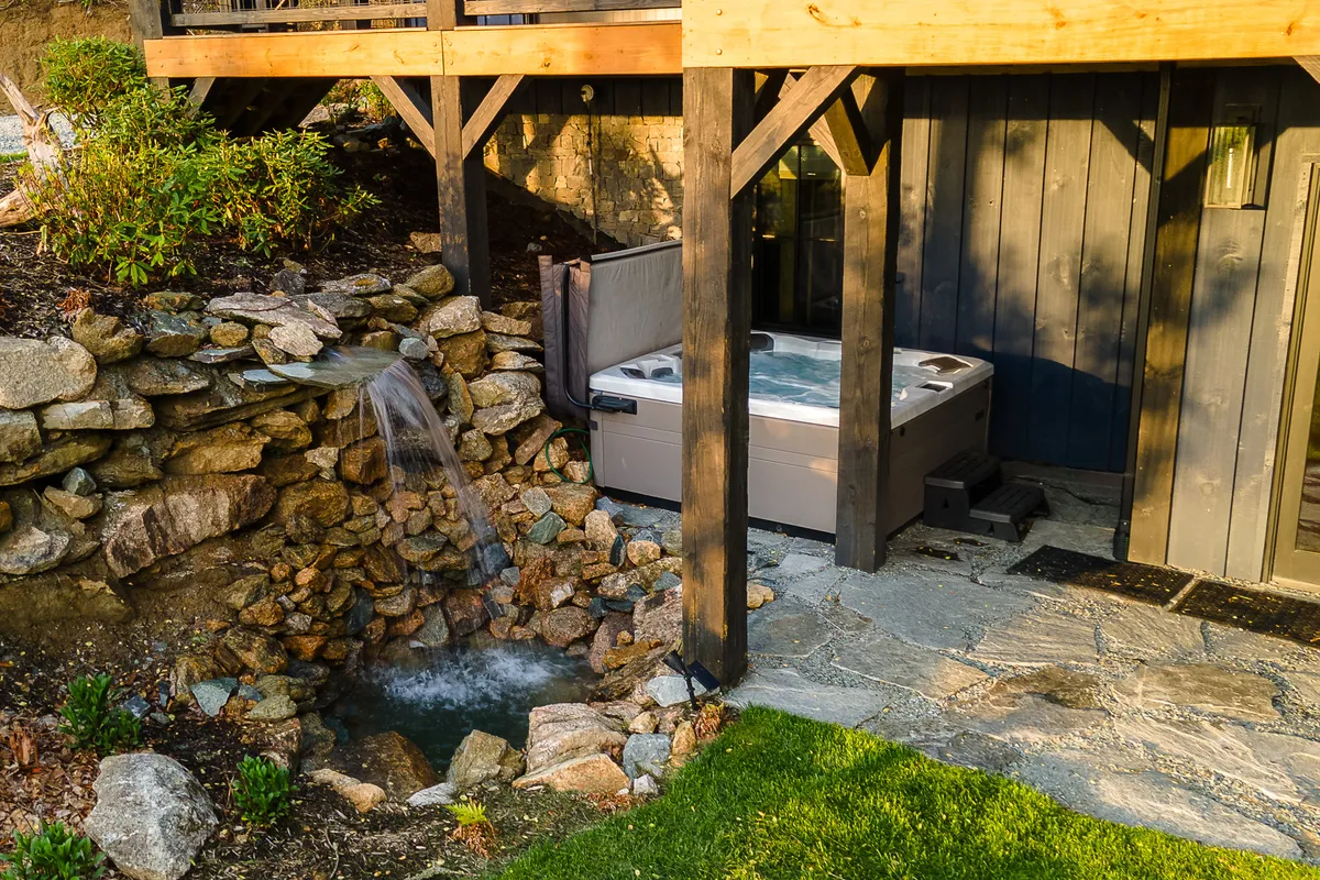 An outdoor scene featuring a private hot tub nestled beside a tranquil waterfall, bordered by a stone wall and surrounded by a lush green lawn. The setting invites relaxation and social gatherings, especially in the evening as the area takes on a serene ambiance.