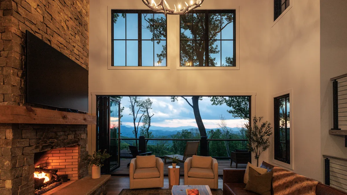 A cozy scene centers around a striking stone fireplace with large windows framing a soft, colorful sunset over distant mountains. Plush seating arrangements invite guests to relax and connect, perfectly blending comfort with scenic grandeur.